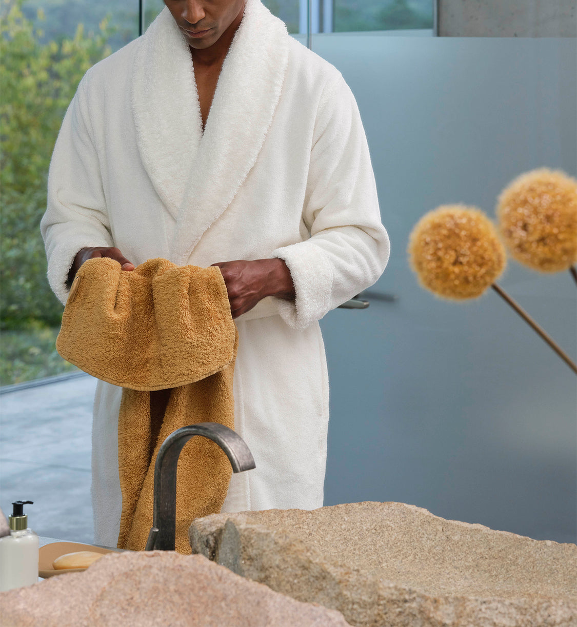 Ein Mann in einem weißen Bademantel hält ein luxuriöses Premium-Handtuch aus feinster ägyptischer Baumwolle in den Händen. Die Szene strahlt Ruhe, Eleganz und die sinnliche Qualität hochwertiger Textilien aus – ein Symbol für pure Entspannung und gepflegten Stil im Badezimmer. Jetzt kostenlose Beratung anfragen – Edle Badmöbel Tiamont.