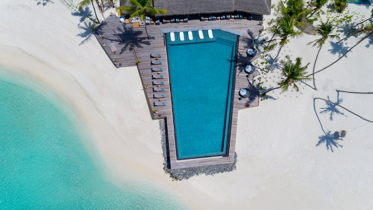 Blick von oben auf einen türkisblauen Pool an einem hellen Sandstrand